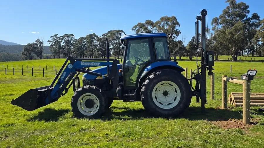 A new farm fence line running over a green rise, post driver at rest beside it.